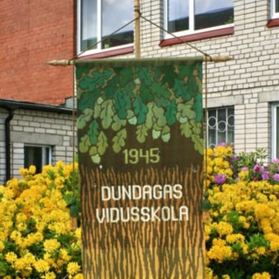 Vertikāls austs karogs ar ozollapu motīvu augšpusē, tekstu 1945 DUNDAGAS VIDUSSKOLA centrā un koka mizas rakstu ar bārkstīm apakšā. Tas ir novietots uz staba ziedu dārzā pie ēkas.