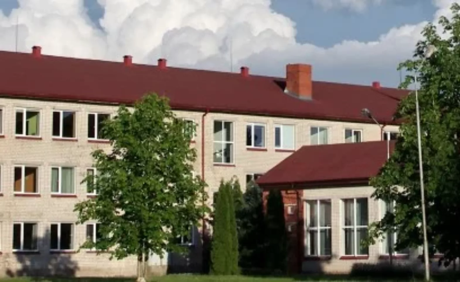 Liela trīsstāvu gaišu ķieģeļu ēka ar sarkanu jumtu, ko ieskauj zaļi koki, zem mākoņainām debesīm.