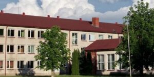 Daudzstāvu ēka ar gaišām ķieģeļu sienām un sarkanu jumtu, iespējams, skola, ar zaļiem kokiem priekšplānā. Labajā pusē redzama ieeja ar savu sarkanu jumtu, pretī mākoņainām debesīm.