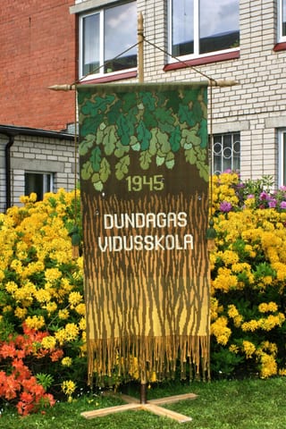 Vertikāls austs karogs ar ozollapu motīvu augšpusē, tekstu 1945 DUNDAGAS VIDUSSKOLA centrā un koka mizas rakstu ar bārkstīm apakšā. Tas ir novietots uz staba ziedu dārzā pie ēkas.