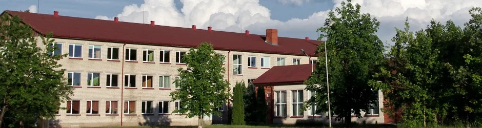 Liela trīsstāvu gaišu ķieģeļu ēka ar sarkanu jumtu, ko ieskauj zaļi koki, zem mākoņainām debesīm.