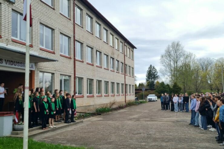 Skolēni un pieaugušie sapulcējušies pie Dundagas vidusskolas ēkas, uz kuras ir uzraksts DUNDAGAS VIDUSSKOLA. Pie skolas ieejas redzams Latvijas karogs un skolēnu grupa zaļos un melnos tērpos uz kāpnēm.