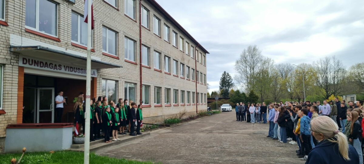 Skolēni un pieaugušie sapulcējušies pie Dundagas vidusskolas ēkas, uz kuras ir uzraksts DUNDAGAS VIDUSSKOLA. Pie skolas ieejas redzams Latvijas karogs un skolēnu grupa zaļos un melnos tērpos uz kāpnēm.
